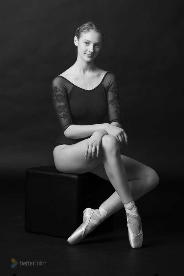 Schwarz-Weiß-Porträt einer jungen Balletttänzerin, die auf einem schwarzen Würfel sitzt. Sie trägt ein schwarzes Balletttrikot mit transparenten Spitzenärmeln und Spitzenschuhe. Mit einem dezenten Lächeln blickt sie direkt in die Kamera. Der dunkle Hintergrund lenkt den Fokus auf ihre elegante, ruhige Haltung und die klassische Tanzkleidung.