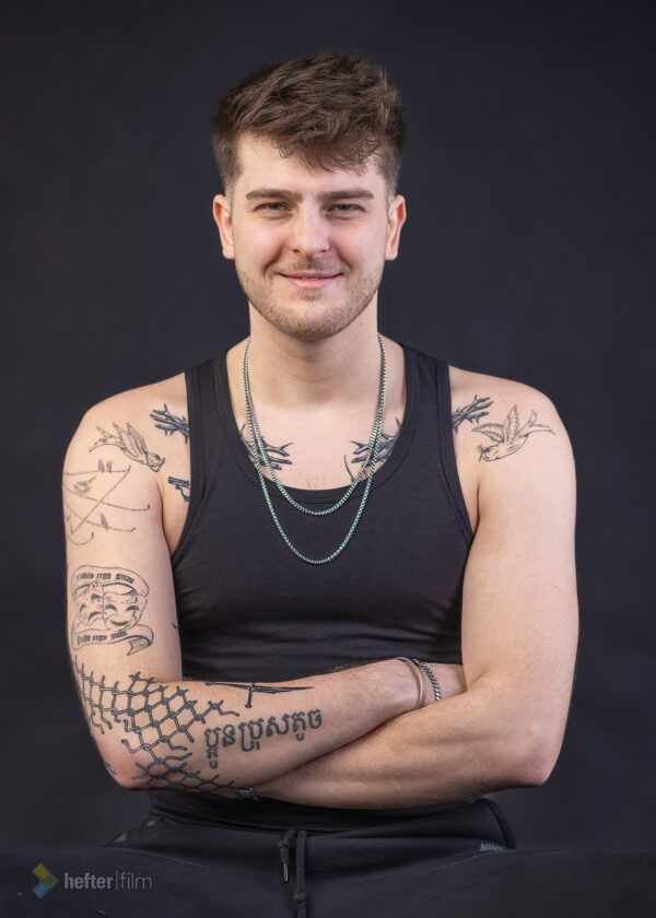 Stani Zechner, Porträt eines jungen Mannes vor schwarzem Hintergrund. Er trägt ein schwarzes ärmelloses Shirt und eine silberfarbene Kette. Mit verschränkten Armen und einem dezenten Lächeln blickt er direkt in die Kamera. Mehrere auffällige Tattoos auf Schultern, Brust und Armen unterstreichen seinen individuellen Stil. Die schlichte Studiobeleuchtung lenkt den Fokus auf seinen freundlichen Gesichtsausdruck und die markante Ausstrahlung.