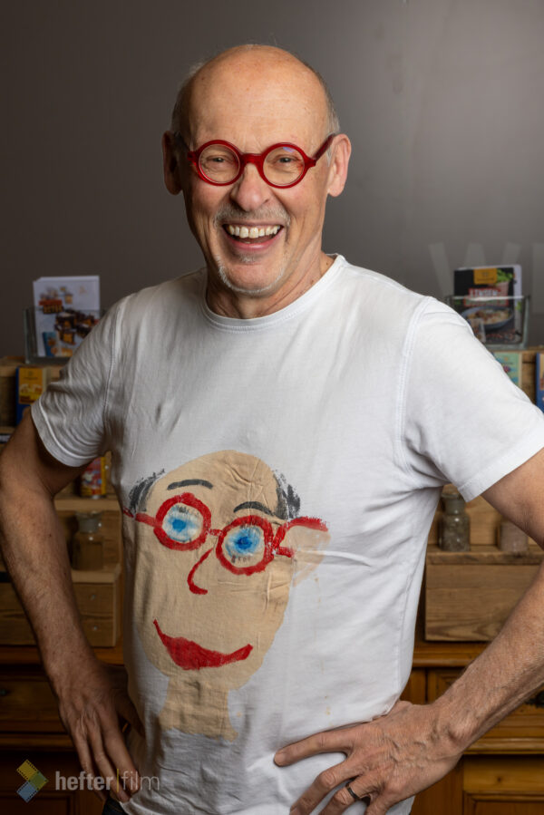 Johannes Gutmann, Gründer und Geschäftsführer von SONNENTOR, lächelt in die Kamera und trägt seine markante rote Rundbrille sowie ein weißes T-Shirt mit einem stilisierten Selbstporträt. Porträtaufnahme des Bio-Pioniers aus Österreich.