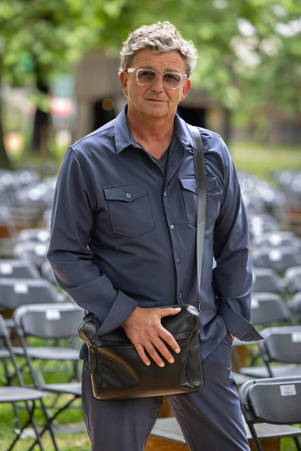 Hans Sigl steht im Freien vor einer Reihe von Sitzplätzen. Er trägt ein dunkelblaues Hemd und eine passende Hose, eine Brille und eine schwarze Umhängetasche.