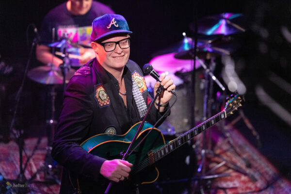 Torsten Goods mit E-Gitarre und Mikrofon auf der Bühne des Porgy & Bess in Wien, begleitet von Schlagzeug im bunten Konzertlicht.