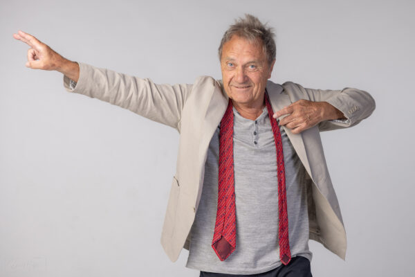 Porträt von Wolfram Berger mit roter Krawatte und ausgestrecktem Arm vor grauem Hintergrund